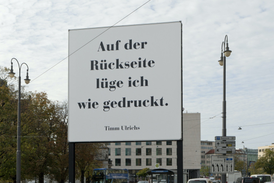 Diagonal view of the billboard. To be read in large black letters on a white background: "Auf der Rückseite lüge ich wie gedruckt. Timm Ulrichs" ("I lie as printed on the reverse. Timm Ulrichs"